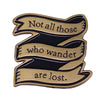A gold-toned enamel pin featuring a black ribbon design with the text Not all those who wander are lost.