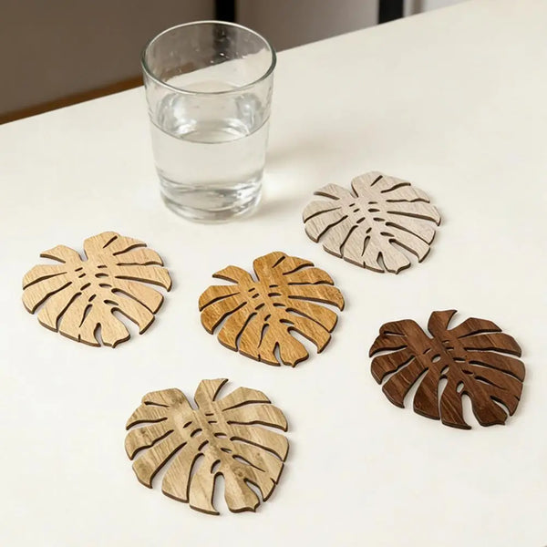 Ein Set aus fünf Holz-Tropikblatt-Untersetzern auf einem weißen Tisch neben einem Glas Wasser.