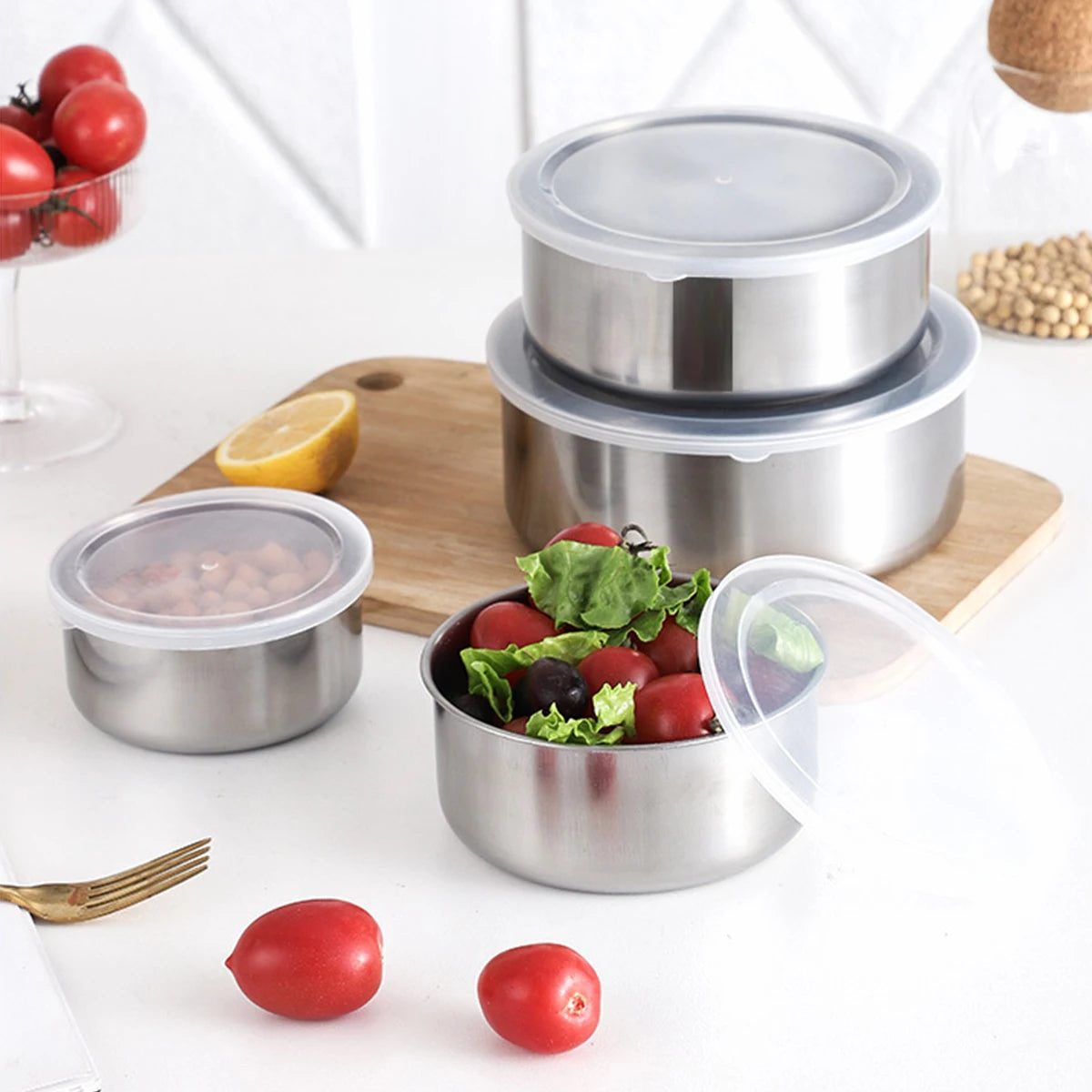 A stainless steel mixing bowl set with airtight lids, various tomatoes, and a lemon on a wooden board.