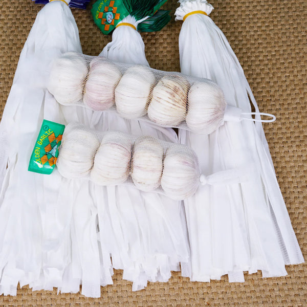 A bundle of white mesh storage bags filled with garlic and ginger are displayed on a woven mat.