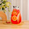 A red glass pitcher filled with sliced citrus fruits and water sits on a wooden cutting board.