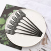 A set of matte black stainless steel dinnerware and cutlery placed on a white plate next to a dark green napkin.