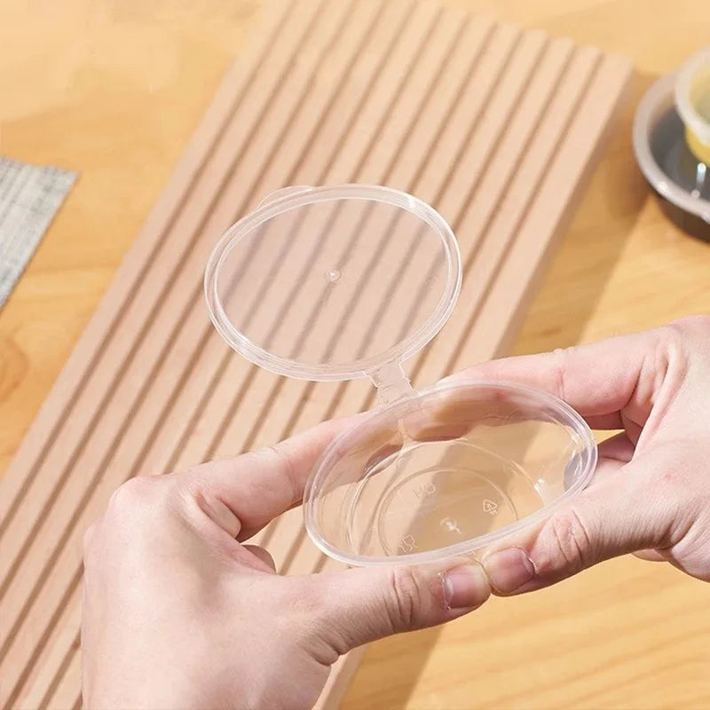 A hand holds a clear, disposable plastic sauce cup for takeaway on a wooden surface.