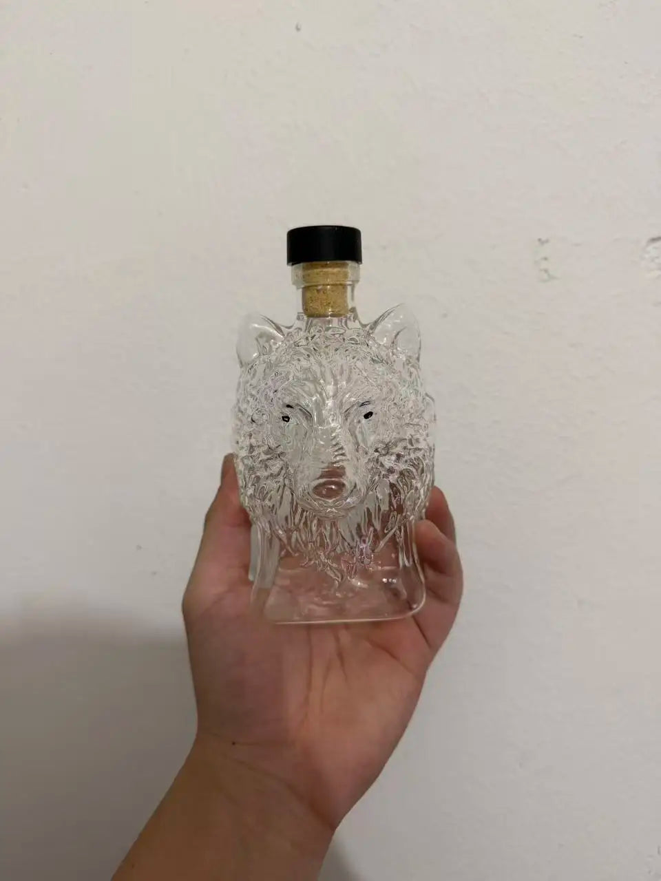 Un decantador de whisky cristalino con forma de cabeza de lobo y un tapón negro, sostenido en una mano.
