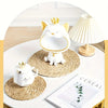 A white ceramic cat figurine with a gold crown sits on a woven rattan placemat next to a white teapot and a lamp.