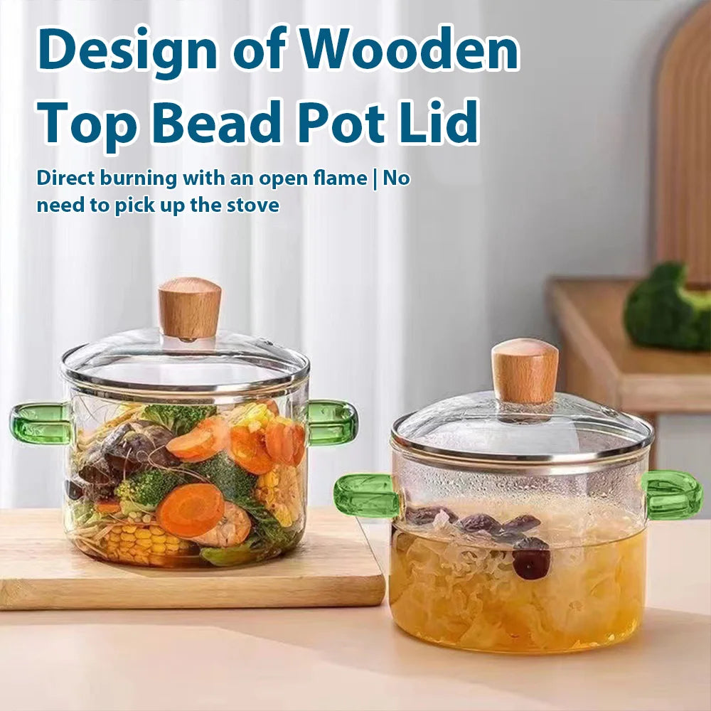 A clear glass soup pot lid with a wooden knob sits on a cutting board, next to a similar pot.