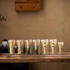 A row of ten glass containers filled with matcha powder and bamboo whisks displayed on a wooden surface.