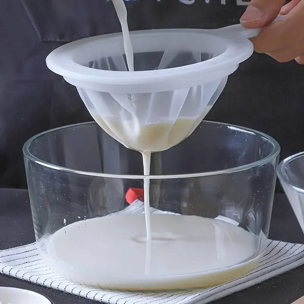 A hand pours white liquid through a multi-mesh fine strainer into a glass bowl.