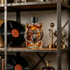 Un decantador de whisky de cristal con forma de cabeza de lobo sobre una estantería de metal rústica junto a un disco de vini