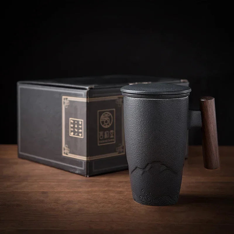 Una taza de té de cerámica negra con tapa junto a su caja rectangular sobre una superficie de madera.