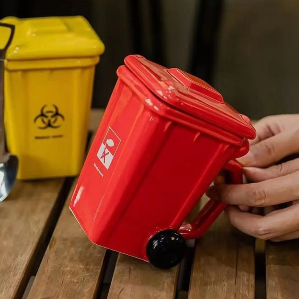 A red novelty trash can is being held by a hand, with a yellow trash can visible in the background.