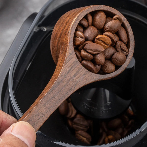 Holzmesslöffel zum Kaffee, gefüllt mit gerösteten Kaffeebohnen, in einem schwarzen Behälter.