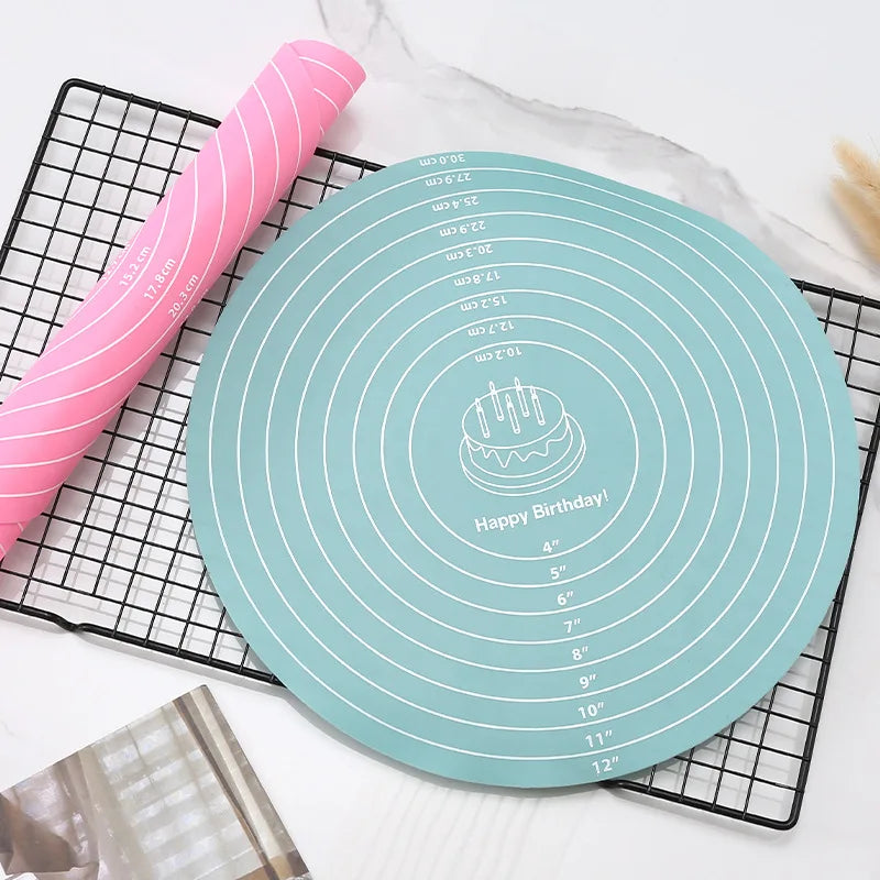 A silicone cake turntable baking mat in light blue is shown on a black wire rack next to a pink silicone cake topper.