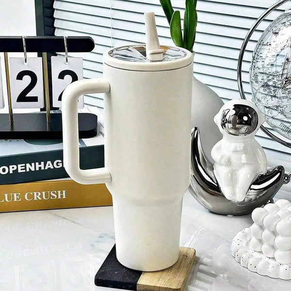 A white stainless steel insulated tumbler with a lid and straw sits on a wooden coaster next to other items.