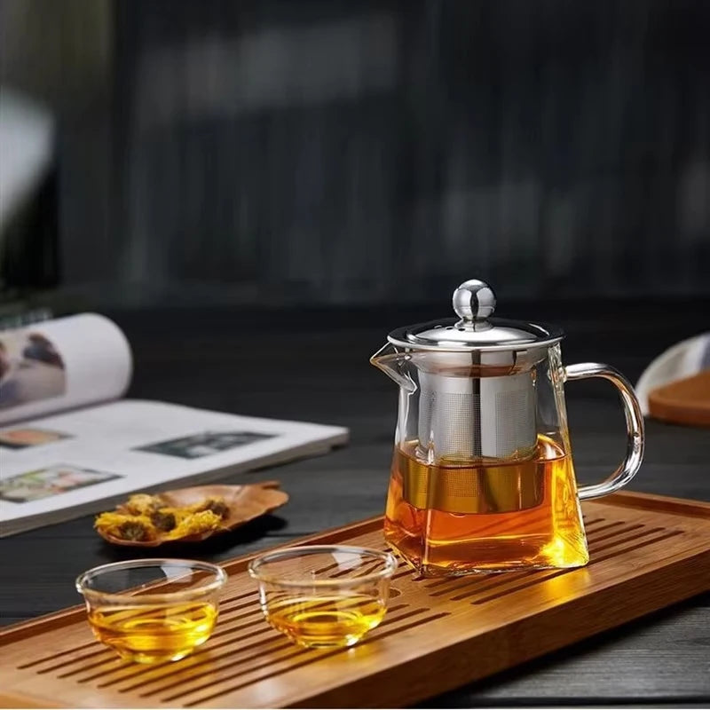 A heated glass teapot with a stainless steel infuser filled with amber tea on a wooden tray.