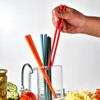 A hand holds a set of red silicone chopsticks over a glass containing green and blue chopsticks.