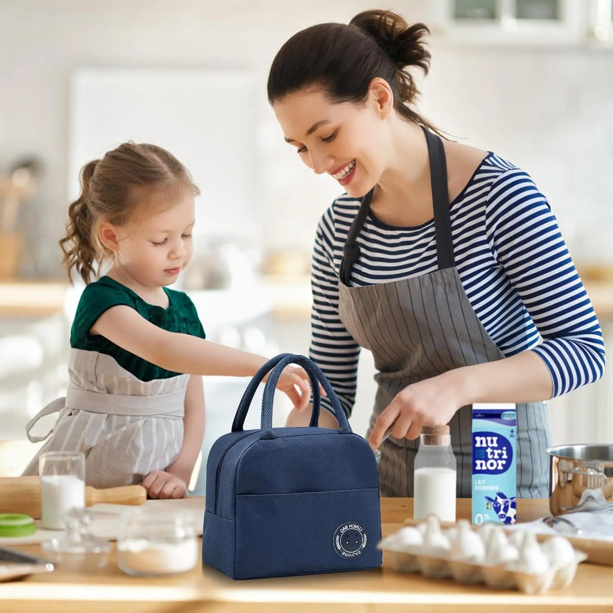 A woman is preparing food in a kitchen with a child watching, next to an insulated blue lunch bag.