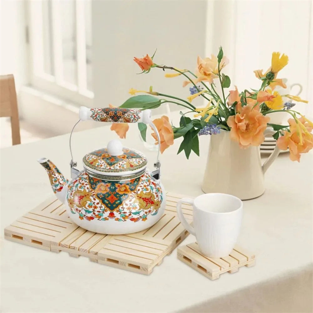 A traditional enamel teapot with floral patterns is displayed on a wooden trivet next to a white cup and flowers.