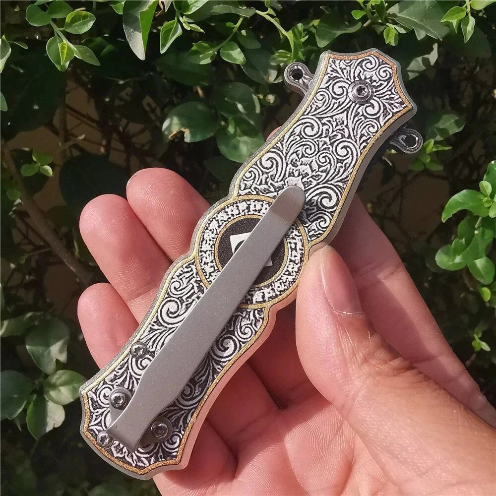 A hand holds a folding utility blade knife with intricate scrollwork decoration against a green leafy background.