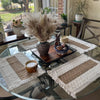 Ein Glas-Tisch präsentiert ein Set Boho-Burlap-Platzsets aus Naturbaumwolle und Fransen, umgeben von getrockneten Gräsern und