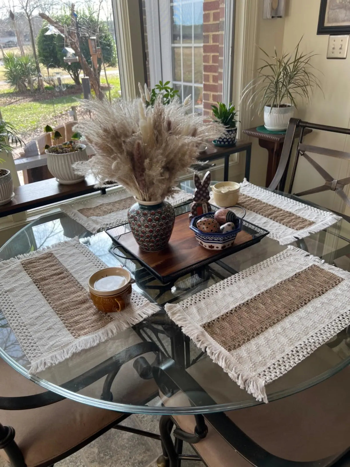 Ein Glas-Tisch präsentiert ein Set Boho-Burlap-Platzsets aus Naturbaumwolle und Fransen, umgeben von getrockneten Gräsern und