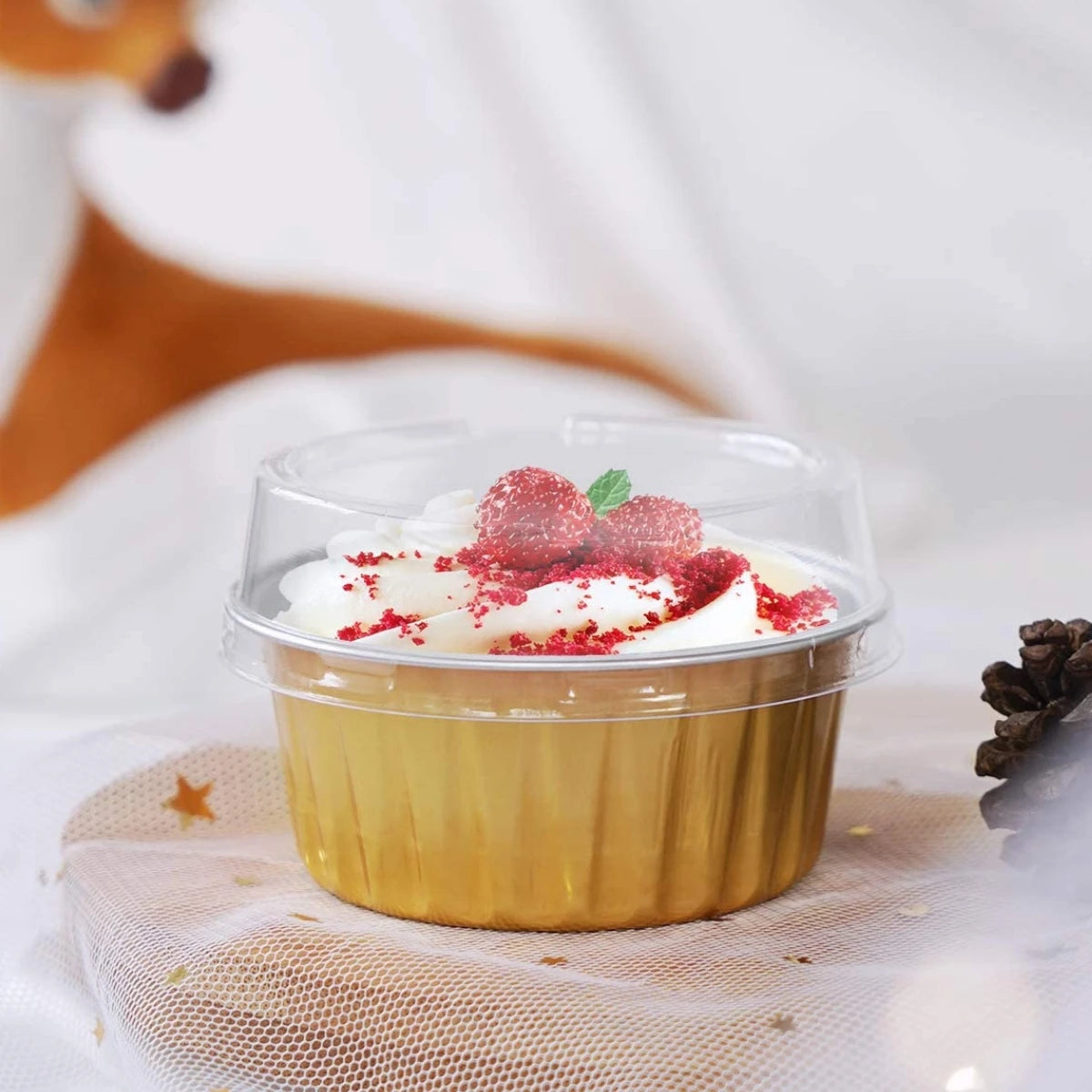 A dessert in a clear aluminum foil baking cup with a lid, topped with whipped cream and red crumbs.