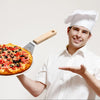 A smiling man wearing a chef's hat holds up a round stainless steel pizza paddle with a wooden handle.