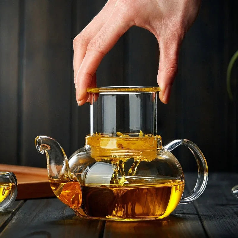 A hand pours amber liquid tea from a high-heat borosilicate glass teapot onto a dark wooden surface.
