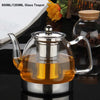A clear glass teapot with a silver lid and handle filled with amber tea, set on a dark surface.
