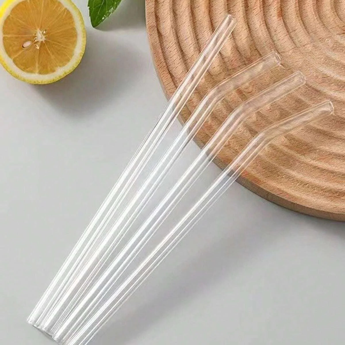 Clear reusable glass smoothie straws with cleaning brushes resting on a wooden board next to a slice of orange.