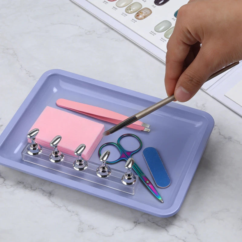 A hand is using a thin metal tool to pick up a pink pencil from a purple tray containing various nail art and dental tools.