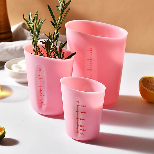 Pinkes Silikon-Messbecher-Set mit doppelter Waage und einer Pflanze in einem Becher, auf einer weißen Oberfläche.