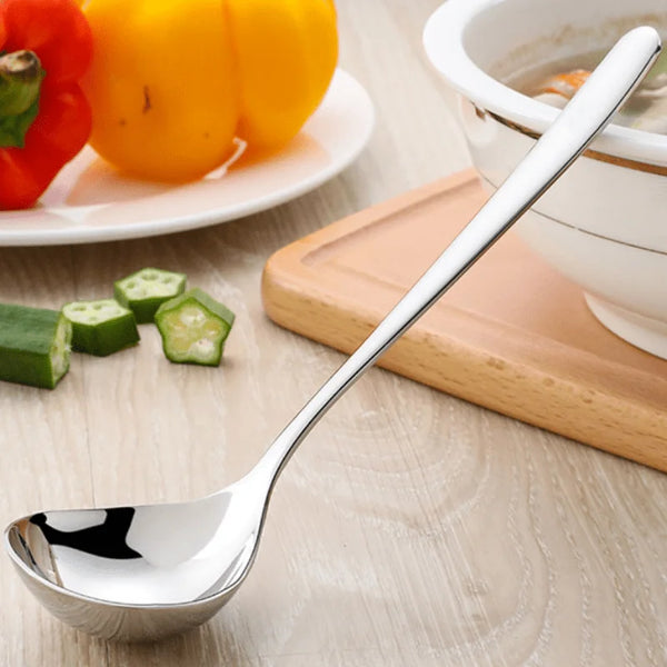 A shiny stainless steel soup ladle with a long ergonomic handle resting on a wooden board next to colorful vegetables.