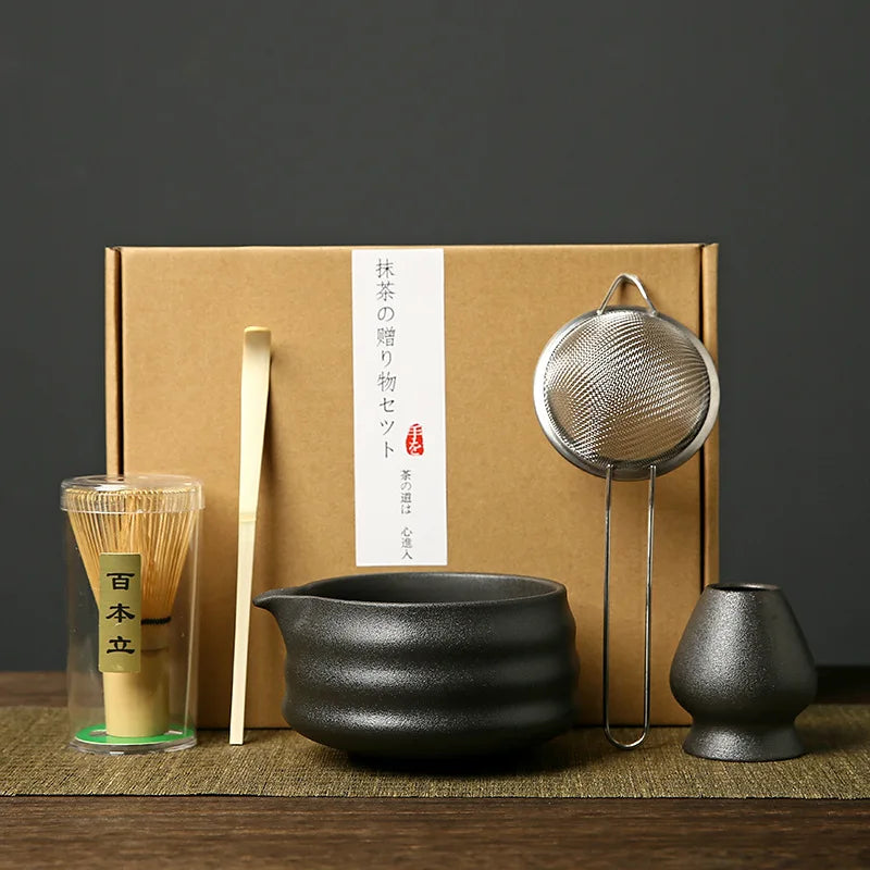 A set of Japanese matcha tools including a bamboo whisk, a metal strainer, and a dark ceramic bowl, presented in a cardboard 