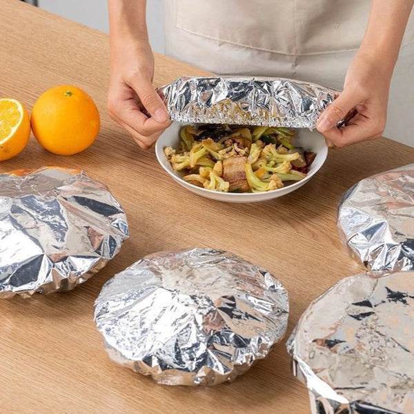 Hände öffnen Aluminiumfolie-Isolierverpackungen mit gekochter Speise auf einem Holztable.