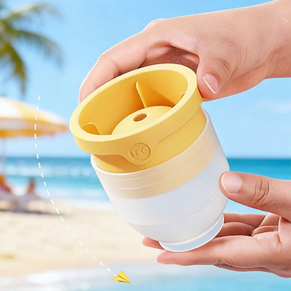 A hand holds a yellow reusable silicone ice ball mold set against a bright blue ocean background.