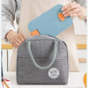 A gray insulated bento lunch bag is shown next to a light blue bag, with a hand holding the blue bag.