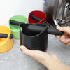 A hand holds a black coffee grounds container with an attached black plunger, next to green and red containers.