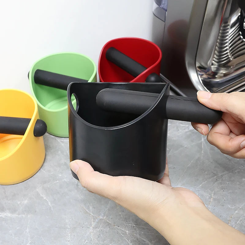 A hand holds a black coffee grounds container with an attached black plunger, next to green and red containers.