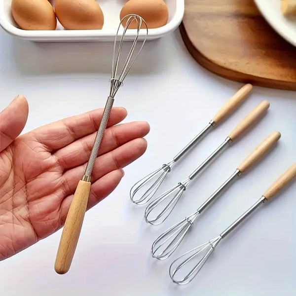 Eine Hand hält ein Mini-Schneebesen-Set aus Edelstahl neben braunen Eiern und einem Holzschneidebrett.