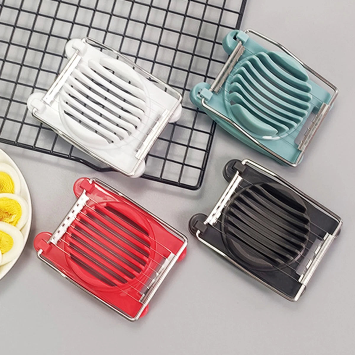 A set of four stainless steel egg slicers and dividers in white, teal, red, and black on a grid background.