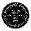 A round black enamel pin featuring white script text that reads Not all those who wander are lost