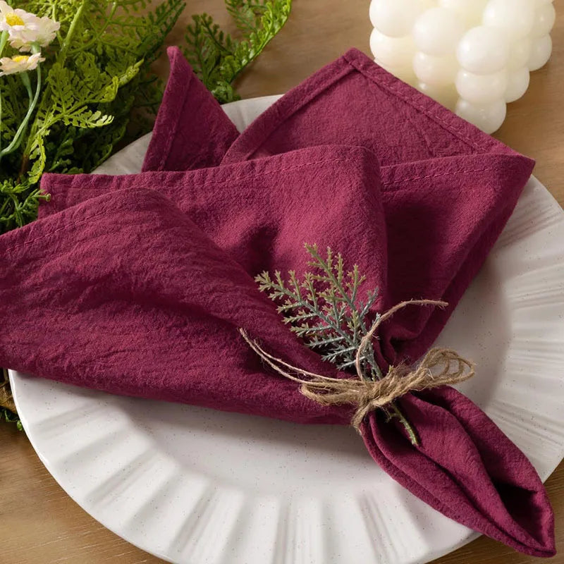 A set of six burgundy cloth napkins is displayed on a white plate with greenery and white grapes.