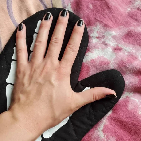 A hand wearing black nail polish is holding a black oven mitt with white accents on a pink fabric background.