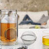 A glass mug filled with amber tea and a stainless steel mesh tea infuser, alongside a small metal sphere and a glass cup of t
