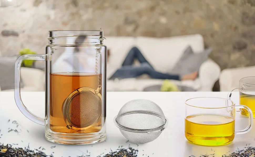 A glass mug filled with amber tea and a stainless steel mesh tea infuser, alongside a small metal sphere and a glass cup of t