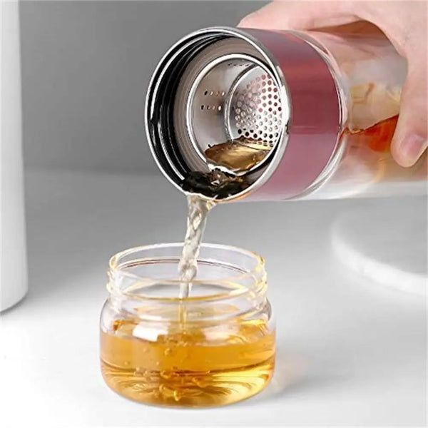 A hand pours amber liquid from a metal tea infuser into a glass jar.