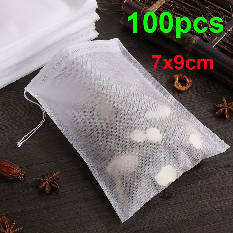 A clear mesh disposable tea filter bag with a drawstring is shown on a dark surface next to dried spices and a wooden tool.