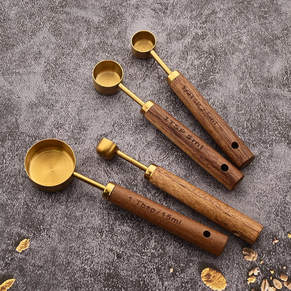 A set of four gold-colored stainless steel measuring spoons with wooden handles resting on a gray surface.