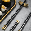A set of five gold style chopsticks resting on a gray surface next to sushi and corn.